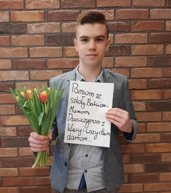 Uczeń SP nr 4 z bukiem tulipanów i planszą z życzeniami: Paniom ze szkoły, babciom, mamom, dziewczynom z klasy i wszystkim damom....